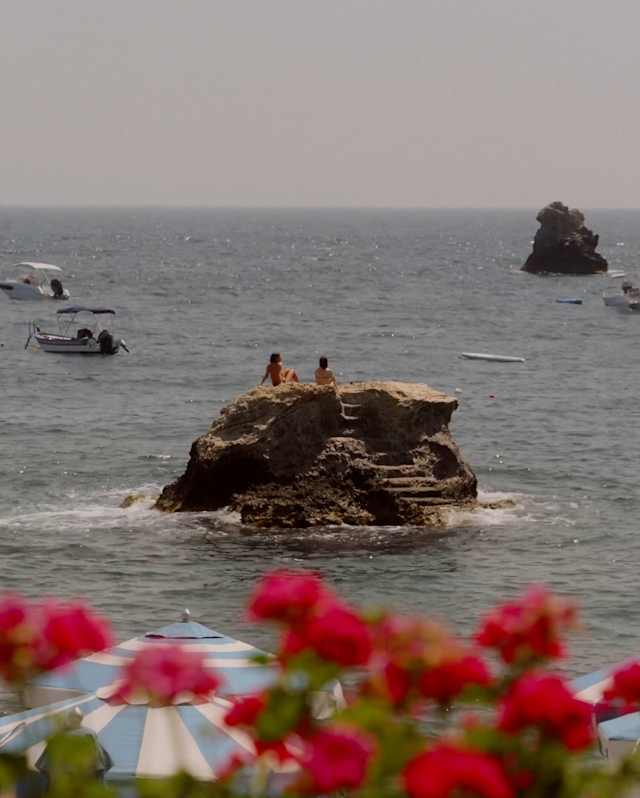 A view of the sea with a few small boats, people sitting on a large rock in the water, and red flowers and striped beach parasols in the foreground.