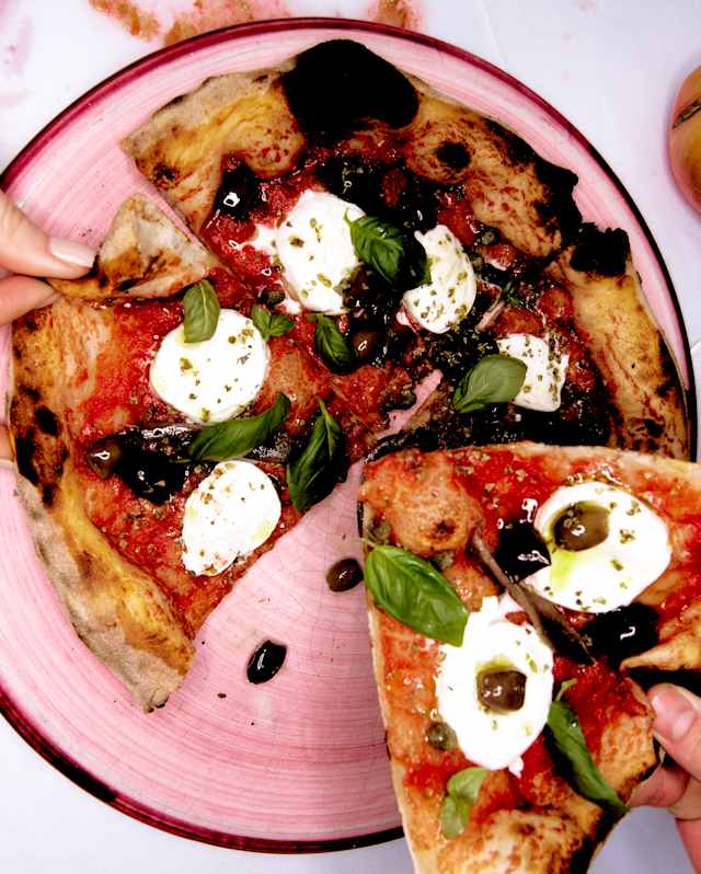 Diners take slices of wood-fired pizza with tomato sauce, fresh mozzarella, basil laves and olives from a pink plate.