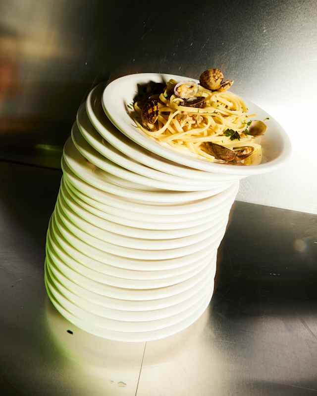 Close-up of a leaning stack of white plates, with the top one carrying a serving of linguine alle vongole, about to fall.