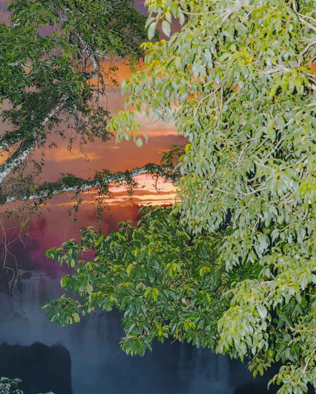 Dense green foliage frames a view of a waterfall with mist rising, set against a vivid sunset sky with shades of orange, pink, and purple in the background.