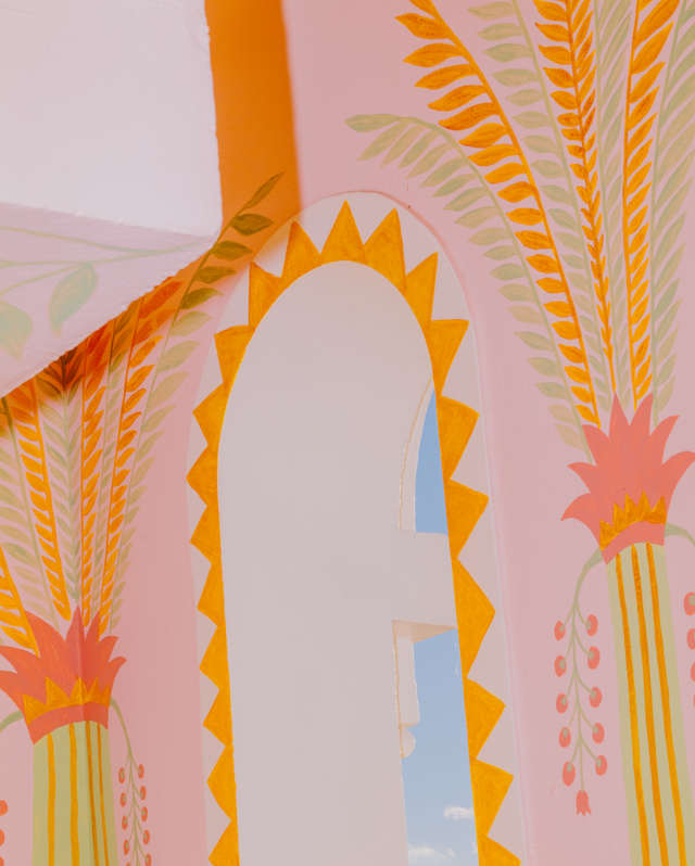 A pastel-coloured archway decorated with yellow zigzag edges and tall palm tree illustrations, revealing a glimpse of blue sky and clouds through the opening.