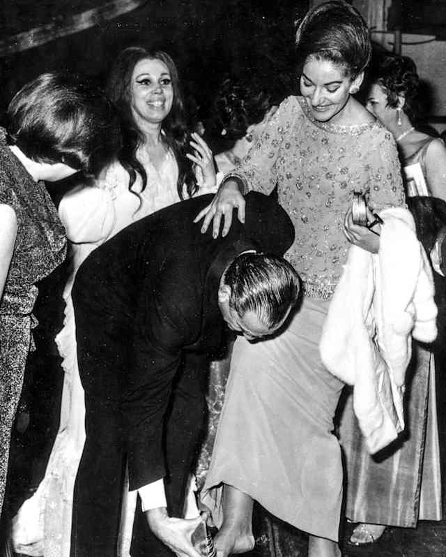 Film director Luchino Visconti stoops to help Maria Callas put her foot in a shoe in a historic black and white photograph.