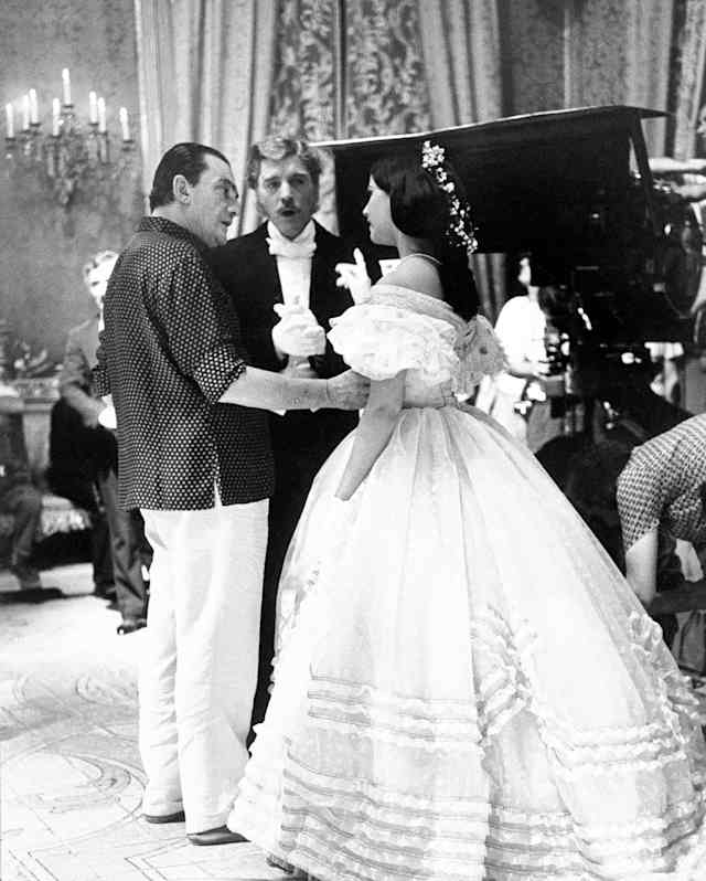 Visconti talks to Claudia Cardinale, who wears a white frilled dress, watched by Burt Lancaster on a film set, in monochrome.