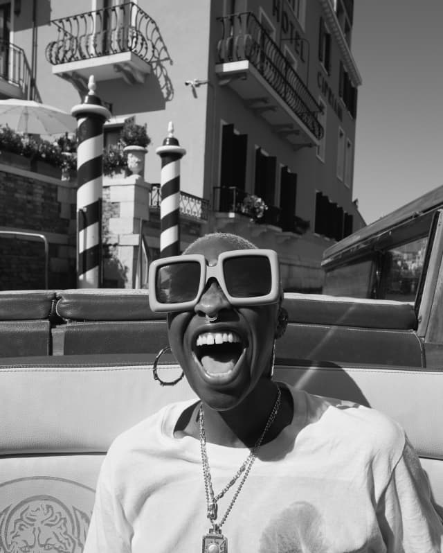 Cynthia Erivo photographed in black and white, laughing in a water taxi, wearing large sunglasses, earrings and a pendant.