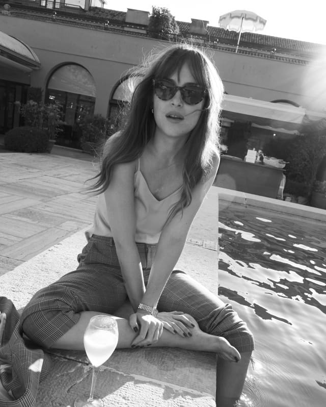Dakota Johnson sits at on the pool edge with sunglasses, a drink, and one foot in the water, photographed in black and white.