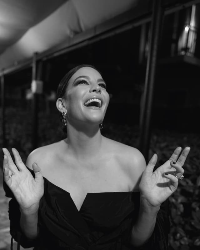 Liv Tyler, in an off-the-shoulder dress Givenchy, raises her hands and laughs, in a black and white capture by Greg Williams.
