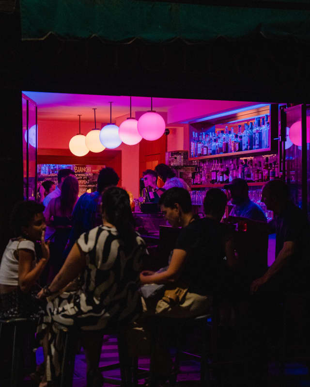 View into a busy bar at night, where barmen take orders beneath a row of five globe lights in pink-purple candy shades.