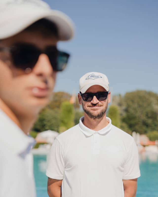 Lifeguard, Andrea Giordano stands by the pool in a white shirt and cap with dark glasses and his arms behind his back.