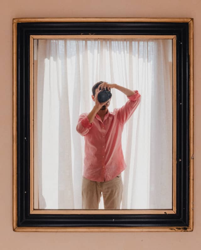 Seen in a mirror's reflection, Content Creation Manager, Marco Valmanara, wearing a pink shirt, holds a camera to his eye.