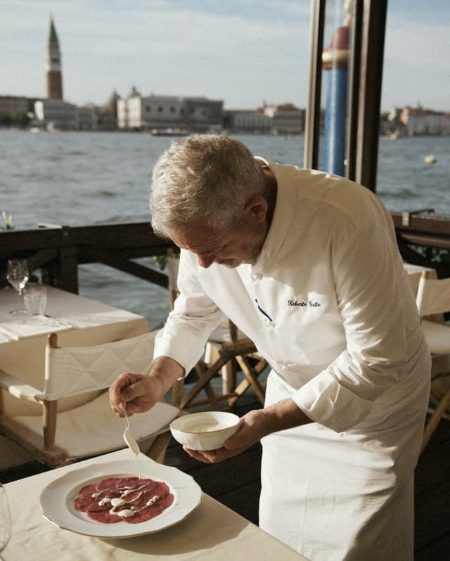 Chef Roberto Gatto, wearing chef's whites, dresses a plate of thinly-sliced bresaola at a Cip's Club waterside dining table.