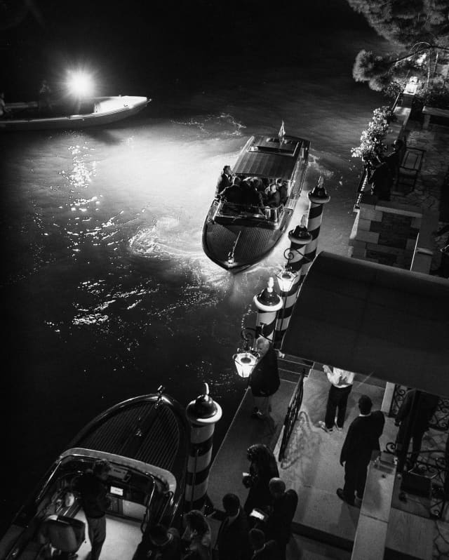 In darkness, hotel staff wait as a late boat pulls up alongside the striped paline posts, guided by lamps, seen from above.
