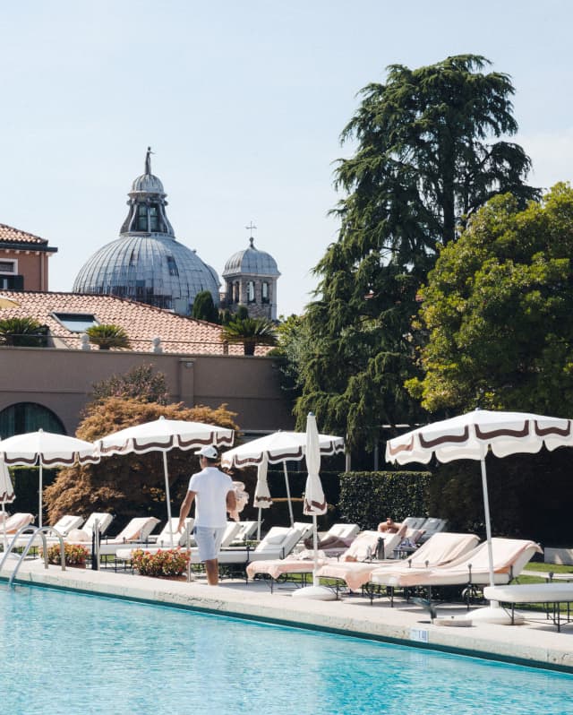 White sun loungers and parasols with brown-trim line the azure swimming pool, with the imposing dome of Le Zitelle behind.