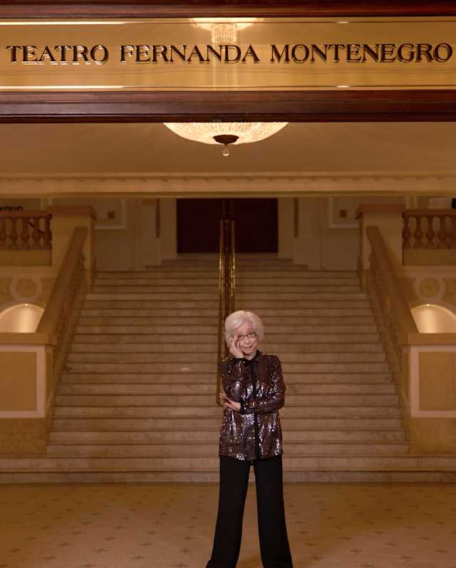 Fernanda Montenegro, with short grey hair and glasses, stands in front of a theatre in her name, wearing a dark sequined top.