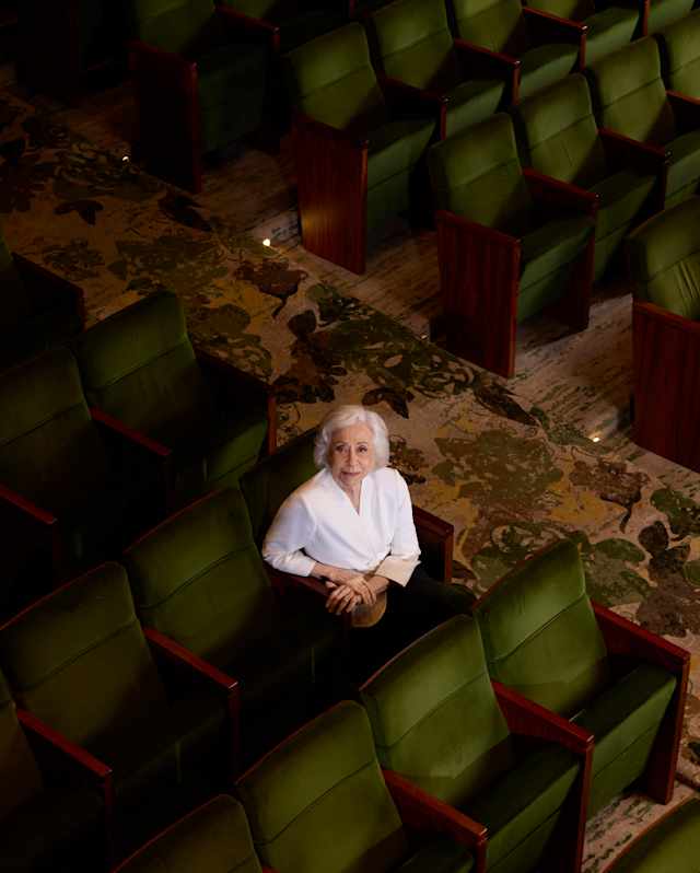 An elderly person with short white hair and a white shirt sits alone in a row of green theatre seats, looking up towards the camera. The surrounding seats are empty.