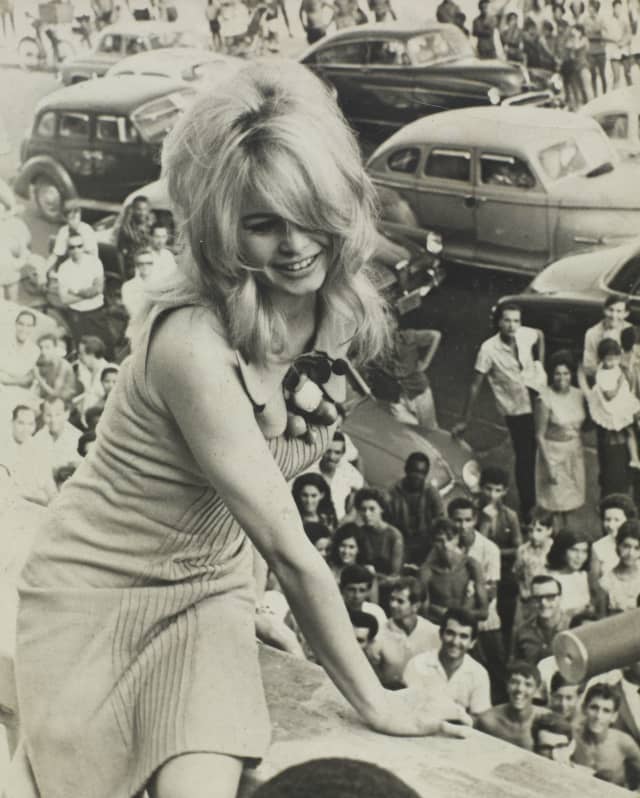 Black and white photo of Brigitte Bardot as she sits on a terrace wall watched by crowds on the street below, taken in 1958.