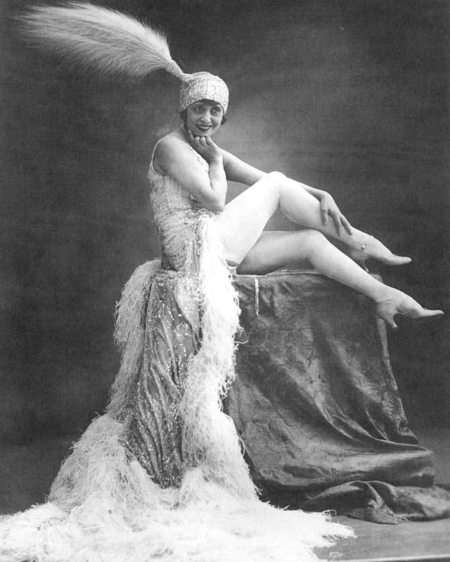 Black and white photograph of Moulin Rouge Burlesque star, Mistinguett, posing on a pedestal in a feather-edged gown and hat.