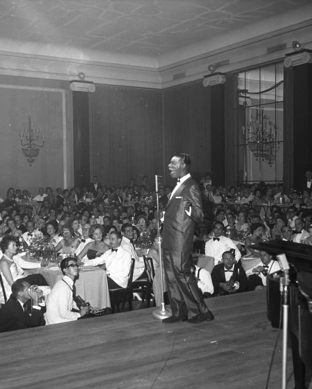 Black and white image of Nat King Cole in a dark suit, presenting on stage to a full audience in the Golden Room in 1959.