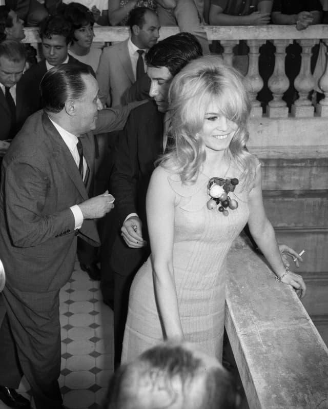 Seen from above, Brigitte Bardot, in a short dress with shaggy blond hair, smiles during a busy outside event at the hotel.