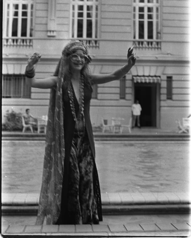 Singer Janis Joplin, wearing round glasses, head scarf, long beads, dress and cloak, puts her hands in the air by the pool.