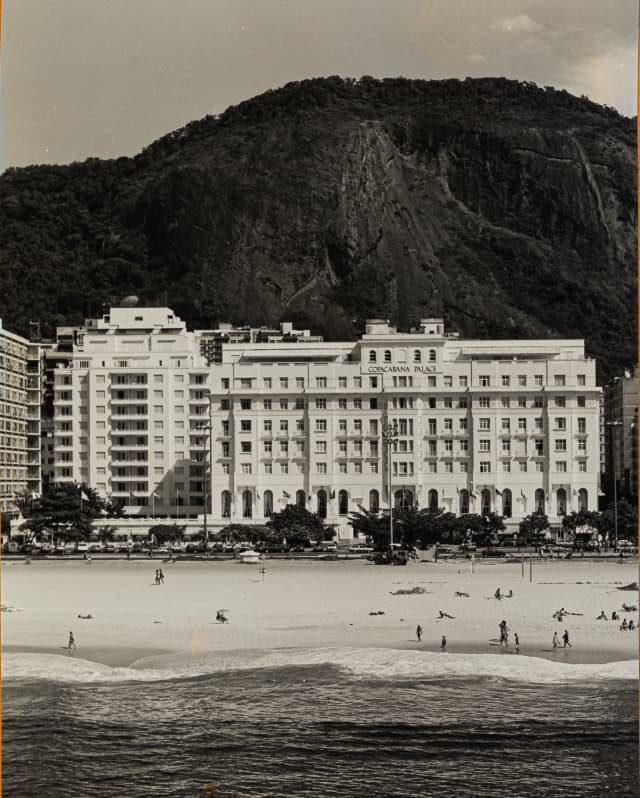 An historic black and white image of Copacabana Palace against the dramatic shape of Morro de São João, seen from the water.