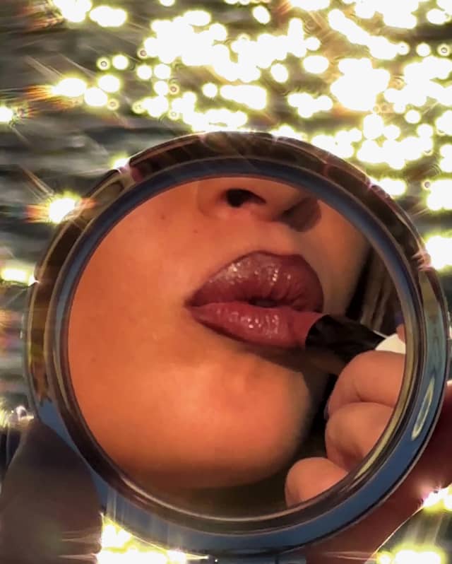 The lower face of a woman applying red lipstick is caught in a round compact mirror, with sea behind, taken by Sam Youkilis.