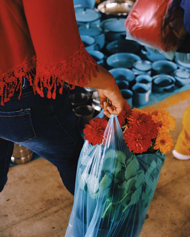 A woman in a tassel-fringed red top carries a plastic bag filled with leafy stems of red and orange marigolds, seen close-up.