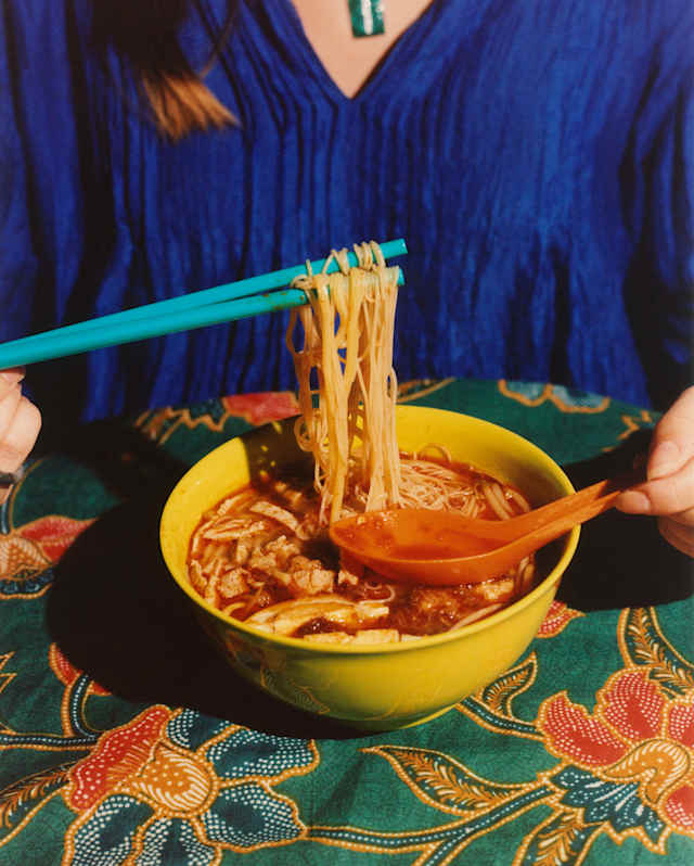A woman in blue eats a noodle broth using chopsticks and a spoon in a picture from the Eat Your Way Through Penang cookbook.