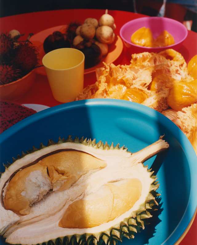 Close-up of a large durian fruit half in a blue bowl on a red table with bright plates of local Malay side dishes.