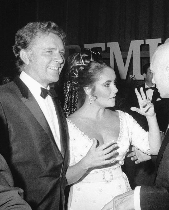 With Richard Burton at her side, Elizabeth Taylor, in a white dress and braided hair, talks to a guest, in black and white.