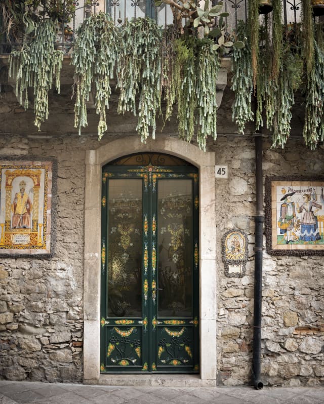 Trailing plants spill from a balcony above the ornate entrance to a ceramics chop with a painted double door and tile-art.