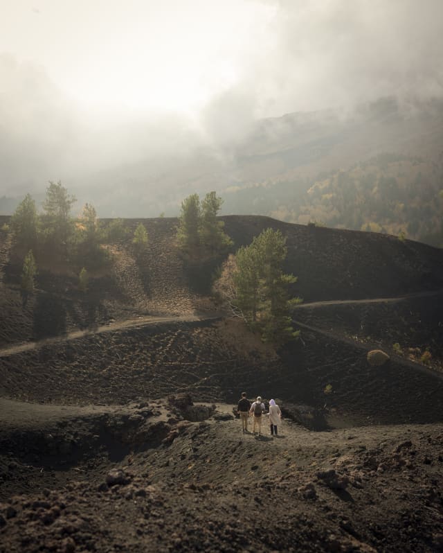 In a low-light image, three walkers hike the undulating, black rocky paths in cloudy Parco dell'Etna, seen from behind.