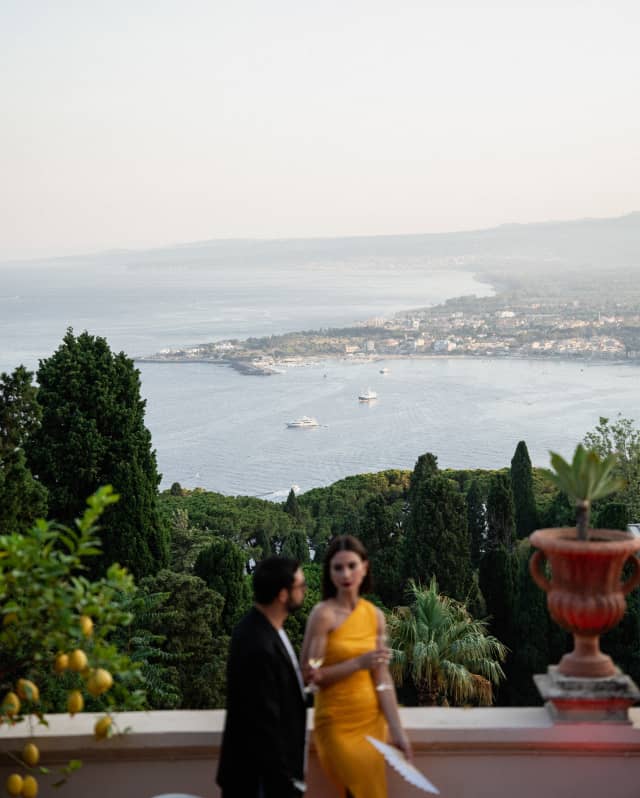 A woman in a yellow dress talks with a man in black jacket on Grand Hotel Timeo's terrace overlooking the Taormina coast.