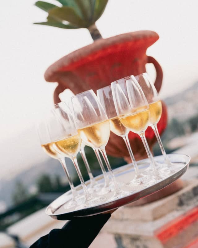 Angled image of eight glasses of chilled sparkling wine on a tray carried by a waiter past a plant potted in a mounted urn.