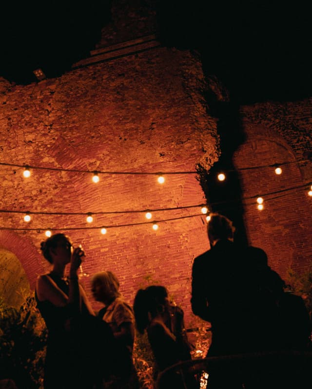 Mingling guests are silhouetted against the reddish glow of an ancient stone wall, illuminated by strings of bulb lights.