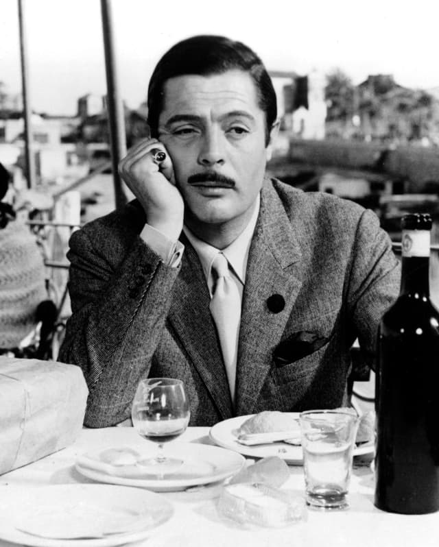 Marcello Mastroianni sits a table with wine overlooking a harbour in Sicily, taken from the film, Divorce Italian Style.