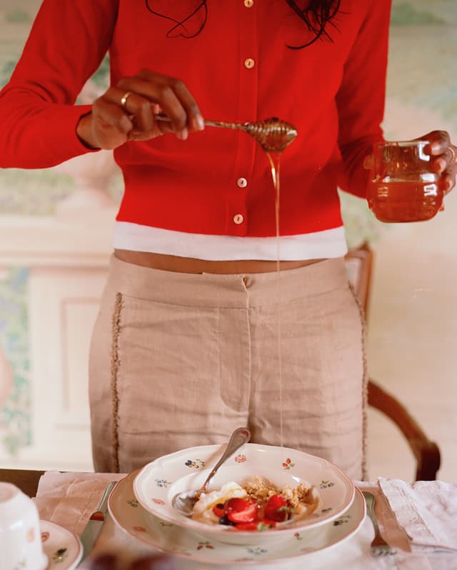 In close-up, a woman in a red cardigan and beige linen trousers holds a honey spool over a bowl of fruit, yoghurt and granola.