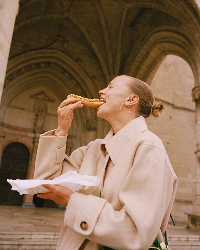 A woman in a beige coat eats a nut-covered chocolate éclair straight from a patisserie paper by the steps of a church.