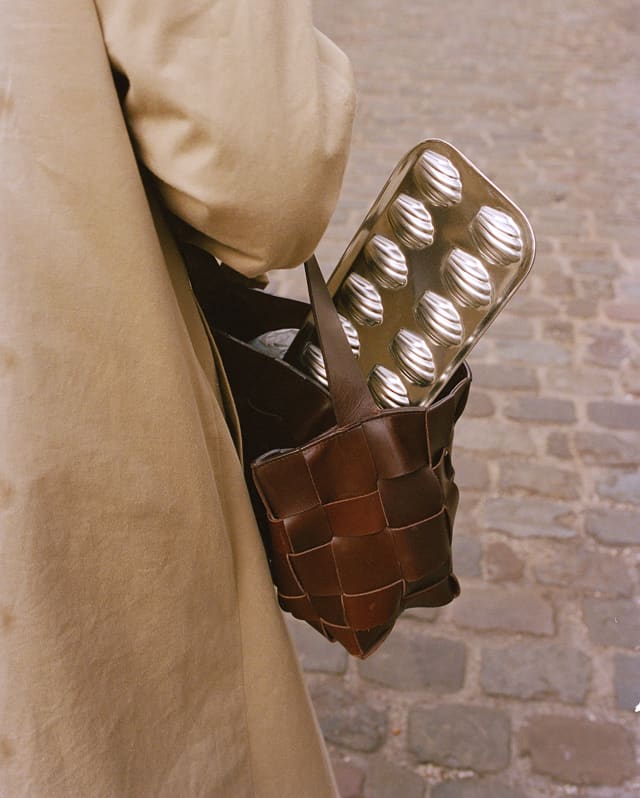 A silver-tone baking tray for making Madeleines juts from the leather bag of a woman in a beige coat, seen from behind.