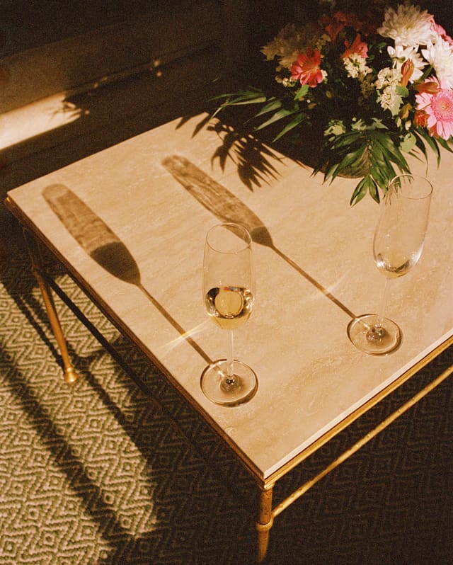 Two Champagne flutes create long shadows across a low table with a flower arrangement, seen from above in evening light.