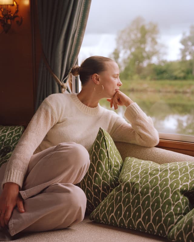A woman in a cream jumper with tied blond hair leans on green Hector-pattern cushions by a window, gazing out at the river.