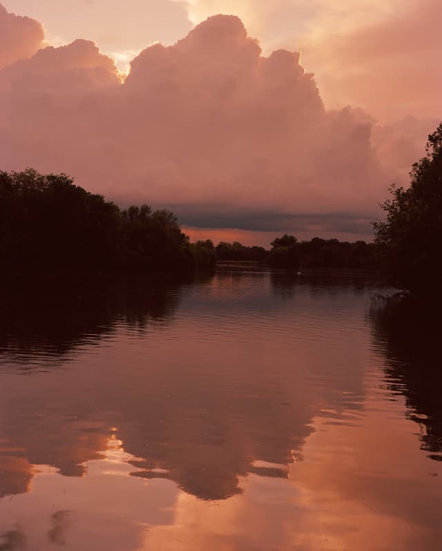 Burgundy clouds bloom in a purpling sky, reflecting the sunset spectacle on the serene waters of tree-lined river below.