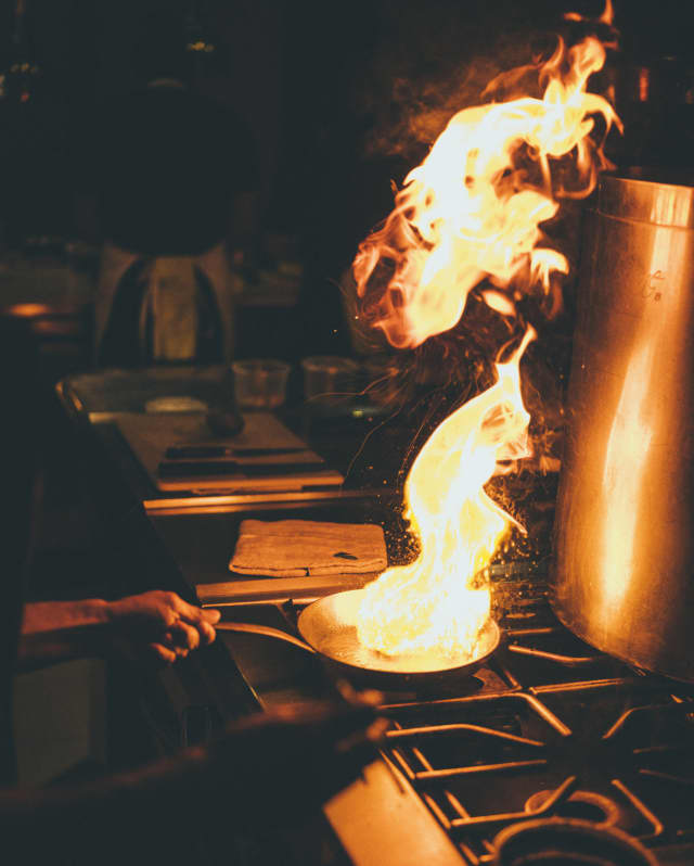 A cook holds a flaming pan on a cooker in a darkened kitchen
