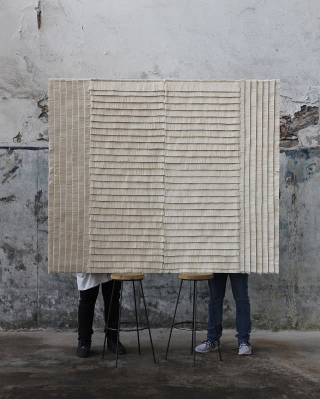 The legs of two men are visible behind a raised Estrella Wall Rug designed by Caralarga using repurposed raw cotton thread.