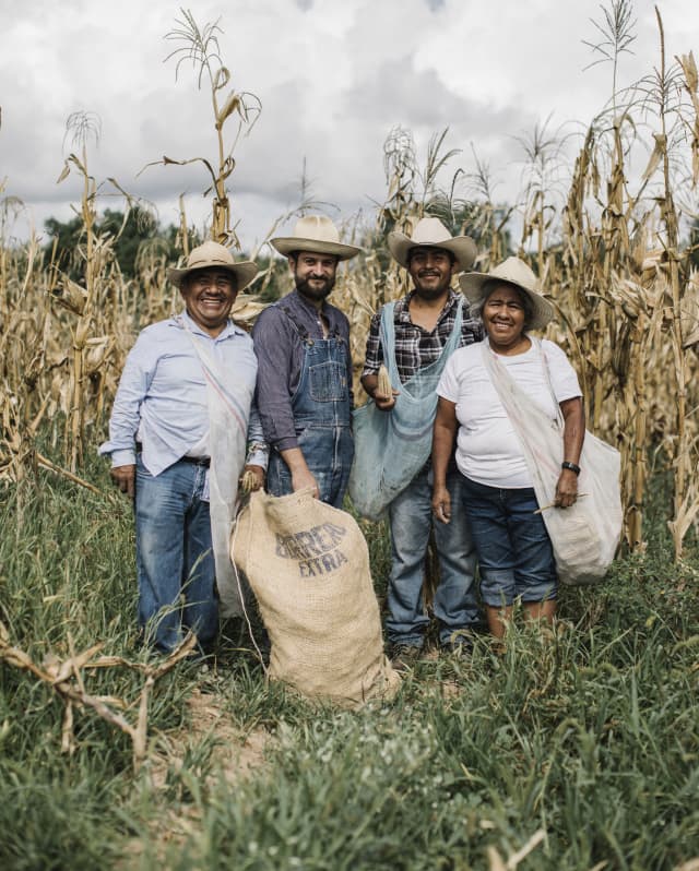 Fernando Laposse, second left, stands in a corn field with three Tonahuixtla farmers, all in white hats, with a sack of husks.