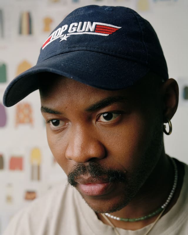 Close-up of designer Lukhanyo Mdingi, with a moustache and earring, wearing a black cap with the red and white Top Gun logo.