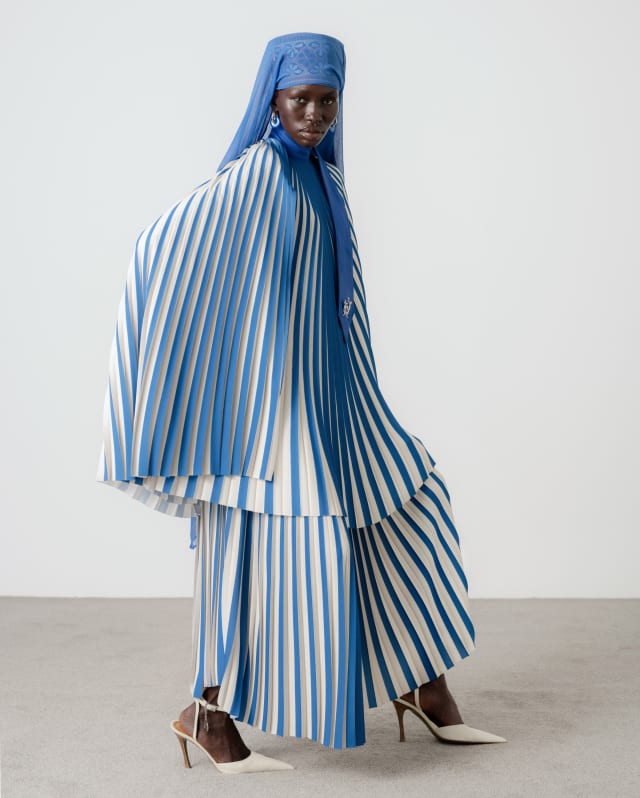A model wears a blue and white striped plissé skirt and cape-like top by Thebe Magugu, with white heels and a blue headdress.