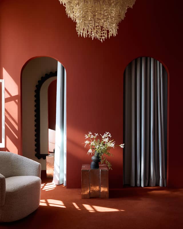 A cosy room with deep red walls and carpet, two arched doorways with curtains, a white textured sofa, a glass side table with a vase of flowers, and a large chandelier overhead. Sunlight streams in through a window.
