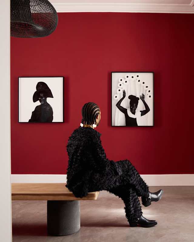 A person in a textured black outfit sits on a wooden bench facing two black-and-white abstract portraits on a deep red wall in a modern, minimalist room with polished concrete floors.