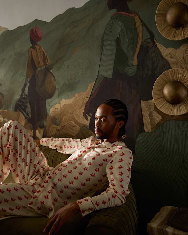 A person with plaited hair, wearing light-coloured clothes with a red pattern, sits on a sofa. Behind them is a mural depicting two figures in traditional attire with a mountainous landscape and decorative wall pieces.