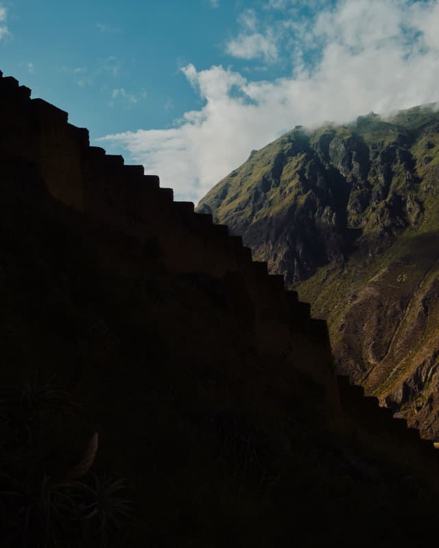 The steps of the Macha Picchu ruins, silhouetted in profile against the green-topped mountains and blue, cloudy sky, behind.